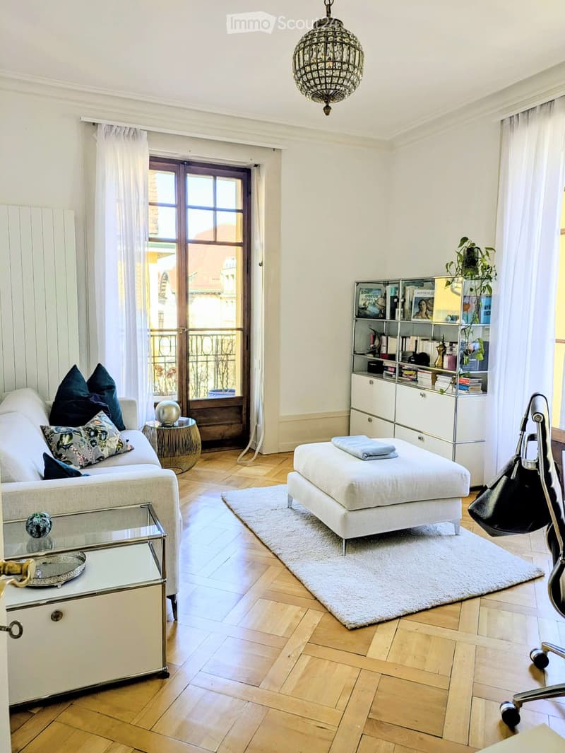 Elegant living room with a white sofa, ottoman, glass coffee table, bookshelf, and a chandelier hanging from the ceiling. The room features wooden flooring and large windows with sheer curtains, leading to a balcony.