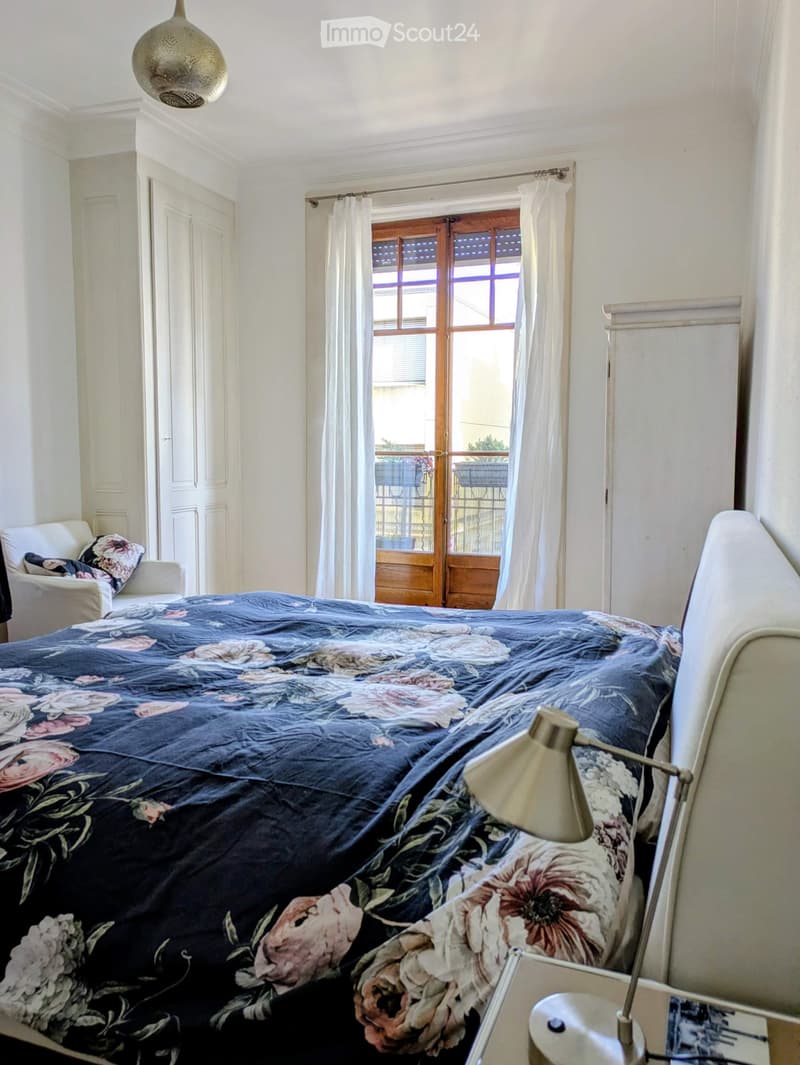 Cozy bedroom with a large bed covered in a floral-patterned duvet, a nightstand with a lamp, a wooden door leading to a balcony, and a small armchair with a patterned pillow.