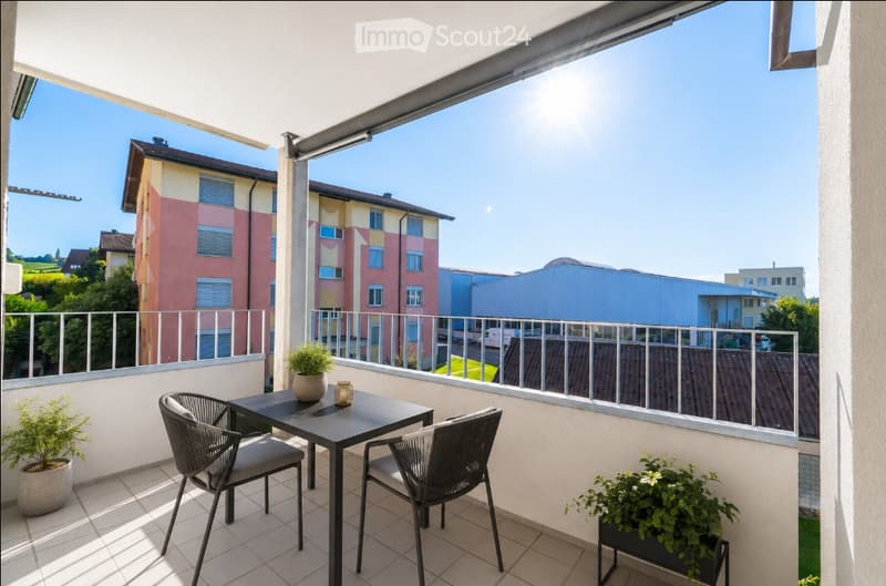 Balcony with a table, two chairs, and potted plants, overlooking a residential area with colorful buildings.