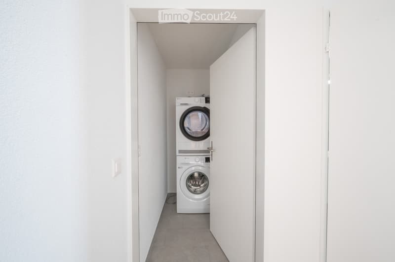 A narrow hallway with white walls and a tiled floor, leading to a room containing a stacked washer and dryer unit.