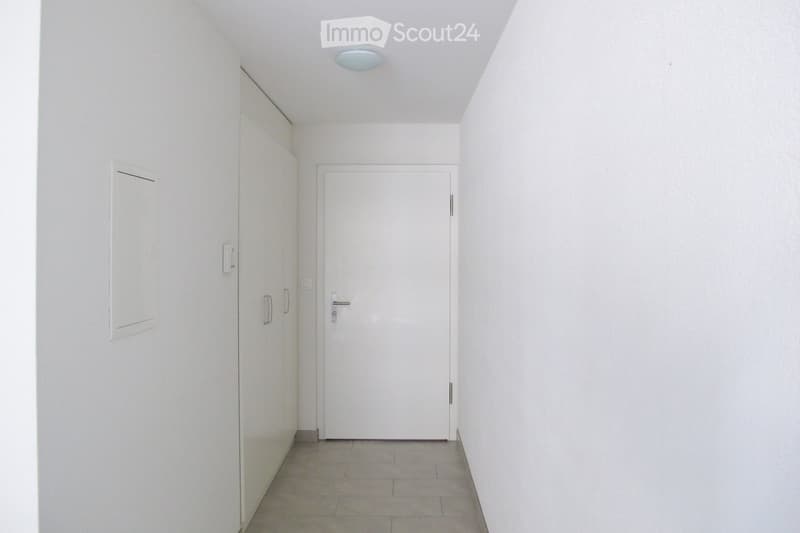 A clean, minimalist hallway with white walls, a tiled floor, and a closed white door at the end. A built-in cabinet is visible on the left side.