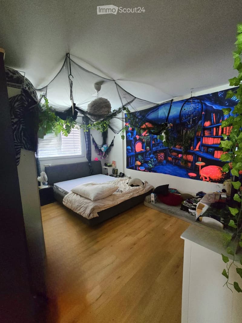 Cozy bedroom with wooden flooring, a large bed with white bedding, a nightstand, a fan, a laptop, a colorful bookshelf mural, and a small plant in the corner.
