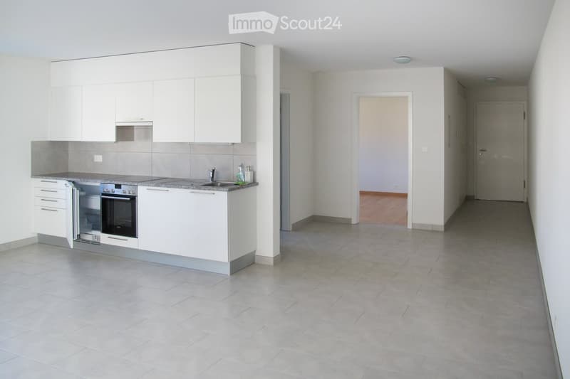 Empty hallway with tiled floor, white walls, and a doorway leading to a kitchen area with white cabinets and stainless steel appliances.
