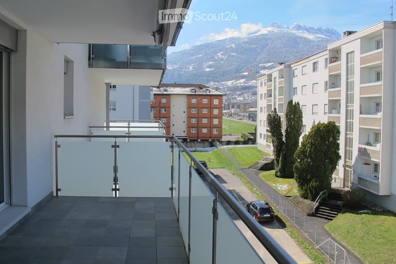 Balcony with modern glass railing, overlooking a landscaped area with trees, parked cars, and distant mountains.