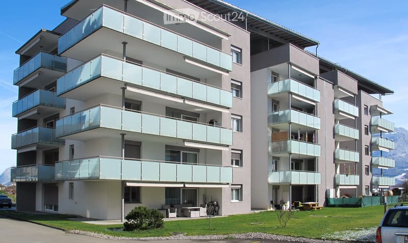 Modern multi-story apartment building with glass balconies, light-colored facade, and surrounding green lawn.