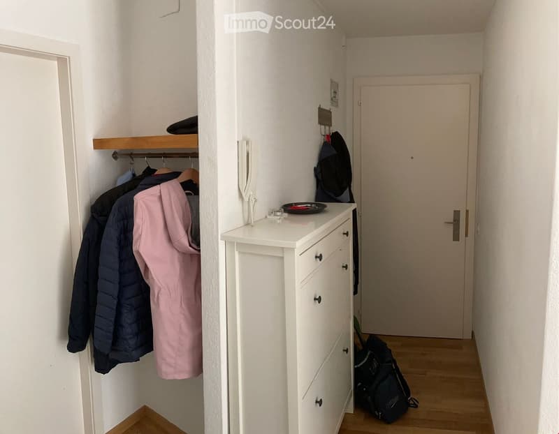 A small room with a white dresser with drawers, a telephone on top, and a jacket hanging in the corner. The floor is wooden and the walls are white.