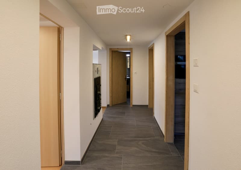 A clean, modern hallway with wooden doors, tiled flooring, and recessed lighting. The walls are painted white, and there are several doors leading to other rooms.