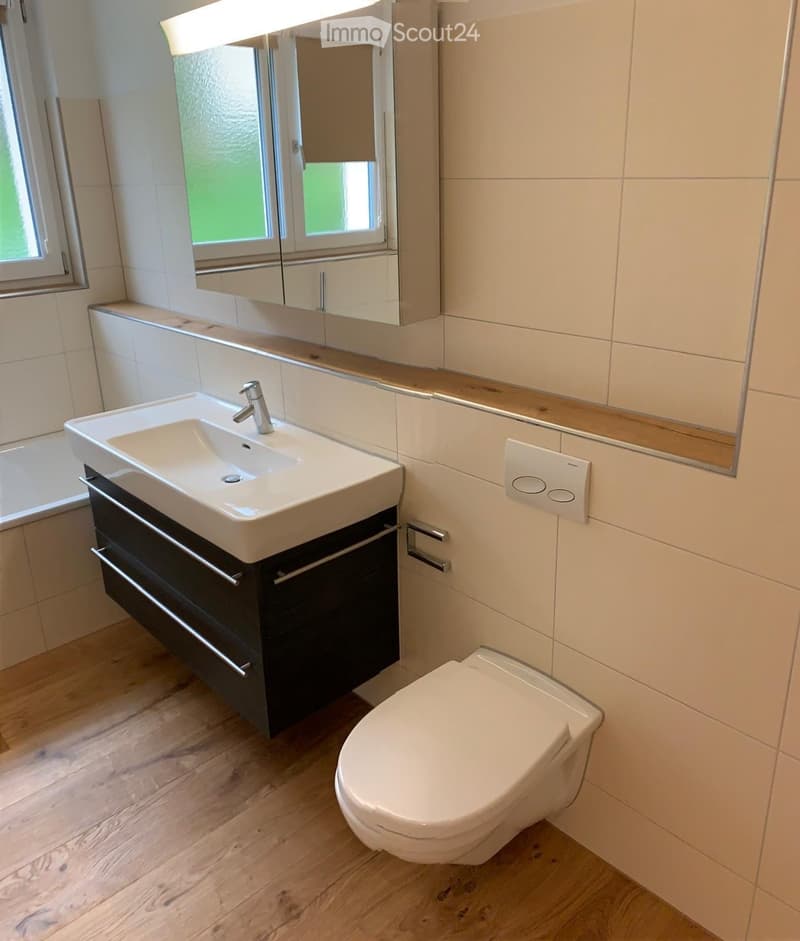 Modern bathroom with a white sink, chrome faucet, mirrored cabinet, and a toilet. Light-colored tiles and wooden flooring.