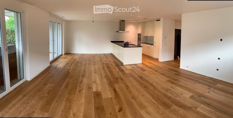 Open-plan space with light wooden flooring, white walls, and a modern white kitchen area featuring cabinets, a countertop, and built-in appliances. Large glass doors lead to an outdoor area.