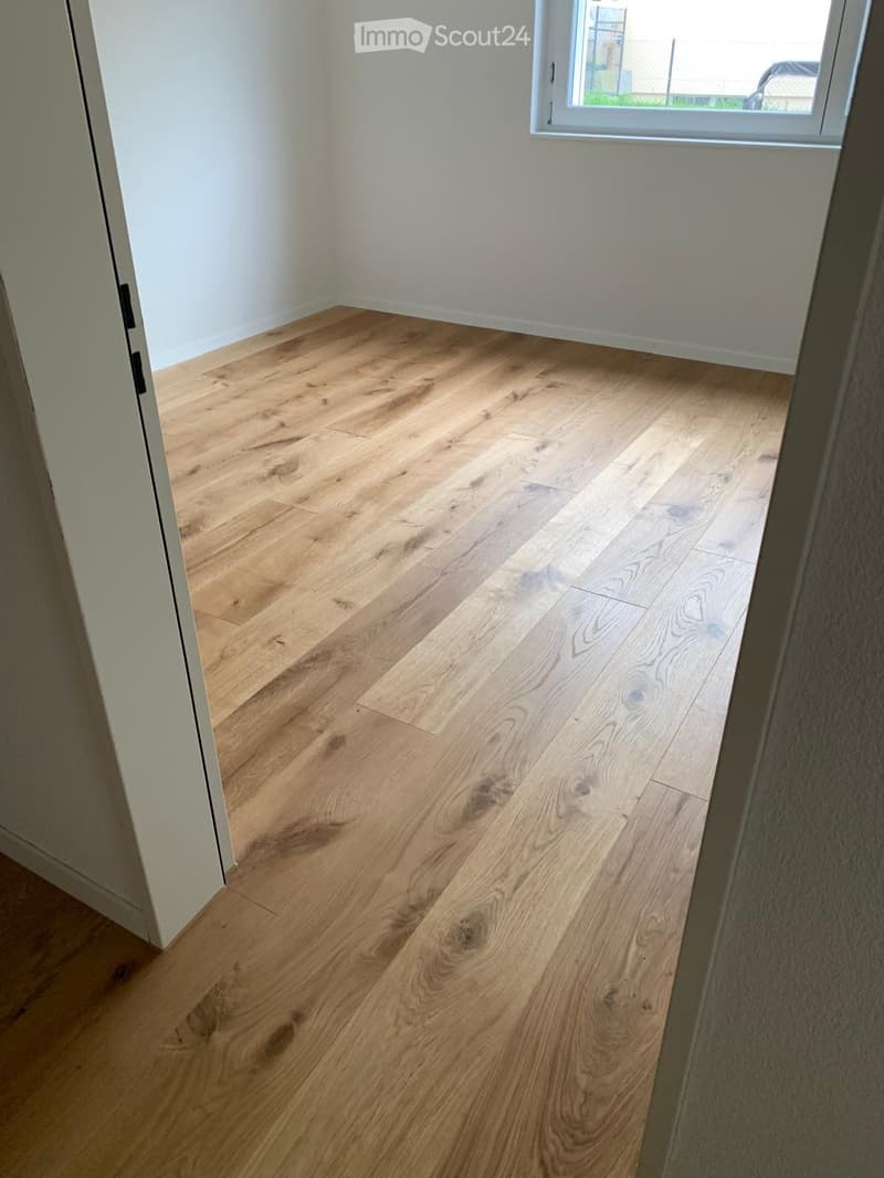 Empty room with light wooden flooring and white walls, featuring a large window on one side.