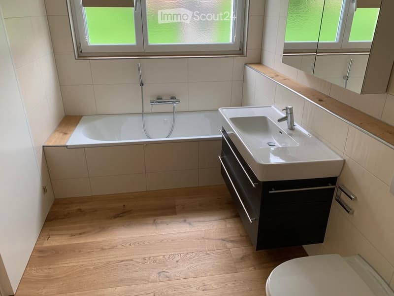 Modern bathroom with a bathtub, sink, and mirror, featuring wooden flooring and tiled walls.