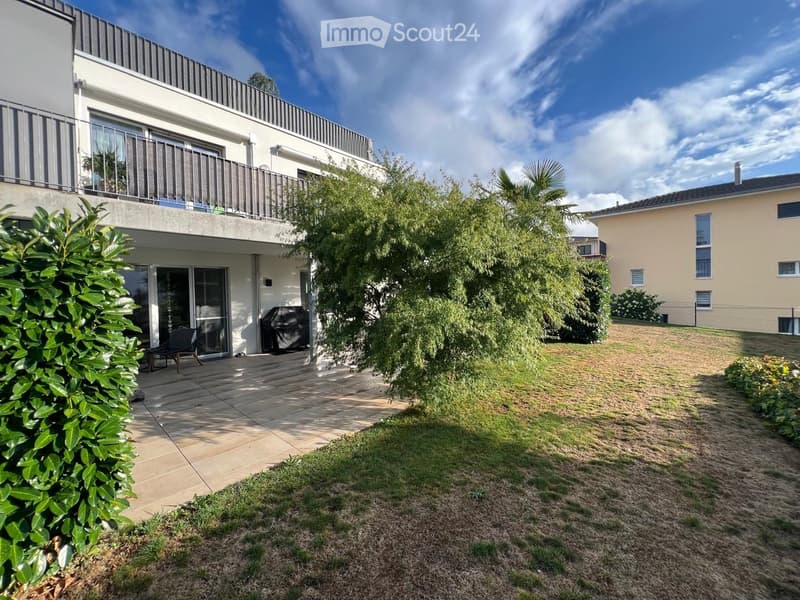 Modern two-story house with a spacious balcony, patio area, and well-maintained garden featuring a lawn, trees, and shrubs.
