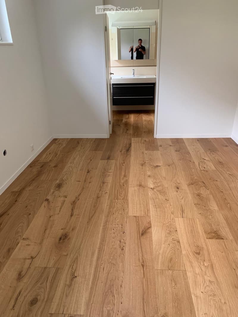 Empty hallway with light wooden flooring and white walls, leading to a bathroom with a sink and mirror.
