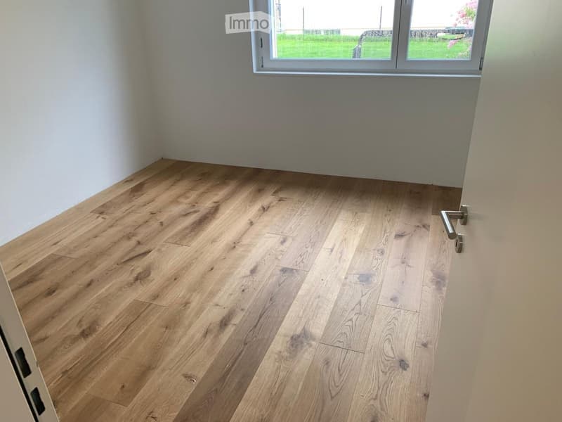 Empty room with light wooden flooring and white walls, featuring a large window with a view of greenery outside.