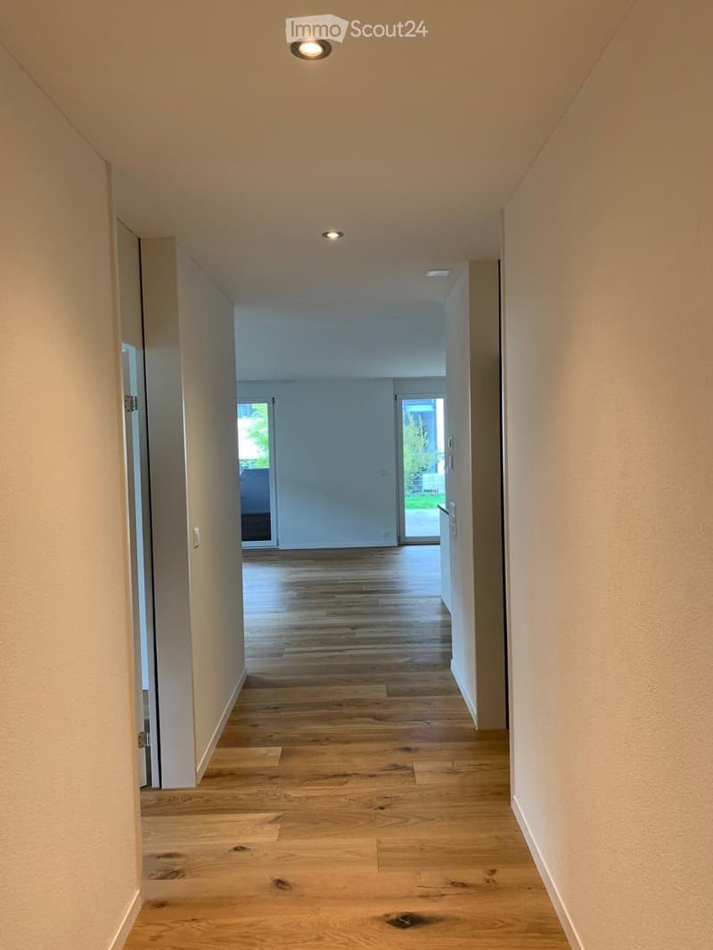 Empty hallway with wooden flooring, white walls, and recessed lighting. Doors lead to other rooms and an outdoor area.