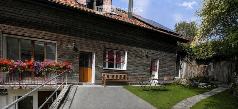 Two-story wooden house with a tiled roof, large windows, and a small garden area with a bench, chairs, and a table.