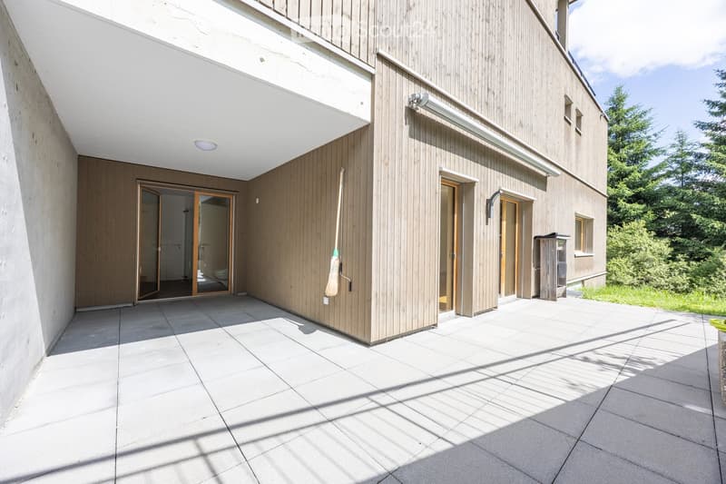 Modern two-story building with wooden siding and concrete elements, featuring a covered entrance area with tiled flooring and multiple doors leading inside.