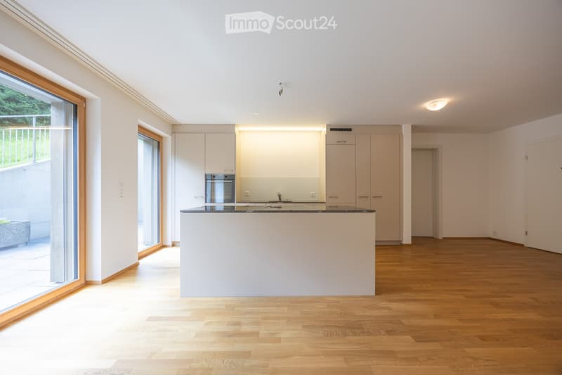 Modern, minimalist kitchen with white cabinetry, stainless steel appliances, and a central island. Large windows provide natural light, and the floor is made of light wood.