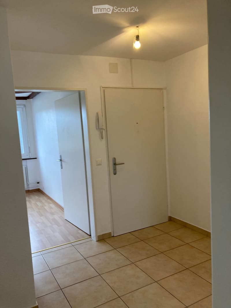 Empty hallway with tiled floor, wooden door, and light fixture. An adjacent room with wooden flooring is visible through an open doorway.
