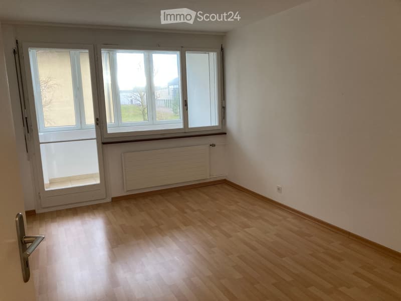 Empty room with wooden flooring, large windows, radiator, and a door handle visible.