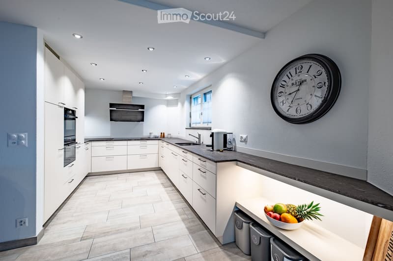 Modern kitchen with white cabinets, grey countertops, built-in oven, and a large window. Features a coffee maker, a bowl of fruit on the island, and a decorative wall clock.