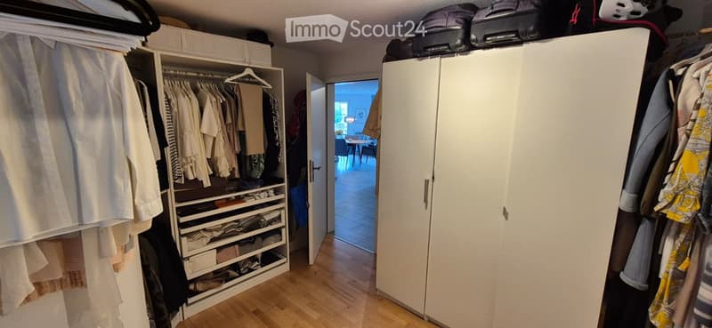 A well-organized walk-in closet with white wardrobes, neatly arranged clothing, and storage compartments. The room features wooden flooring and an open doorway leading to another space.