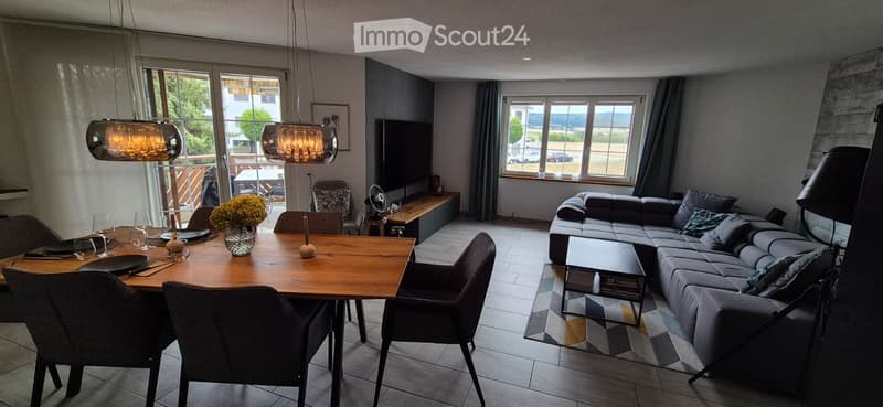 Modern living and dining area with a grey sectional sofa, wooden dining table, pendant lights, and large windows offering a view of the outdoors.