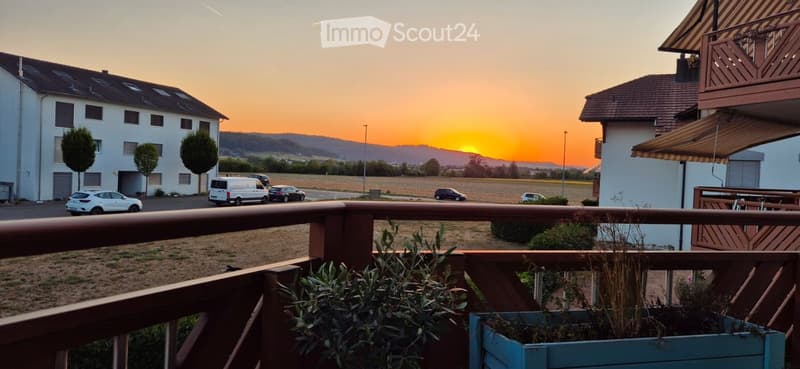 Balcony overlooking a residential area with parked cars, buildings, and a sunset view over a field and distant hills.