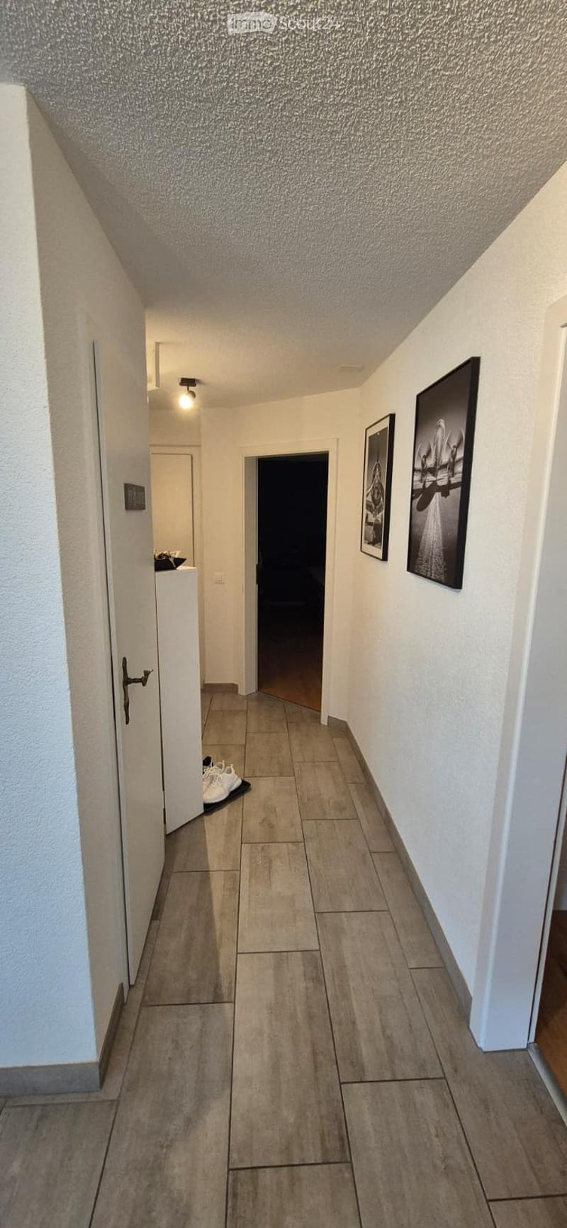A modern hallway with tiled flooring, white walls, and two framed pictures. A small shoe rack with white shoes is visible near the left door.