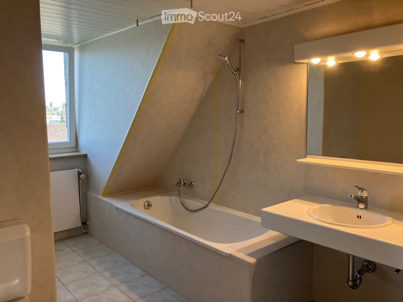 Modern bathroom with a bathtub, sink, mirror, and a window with a view. Features a sleek design with light-colored tiles and a radiator.