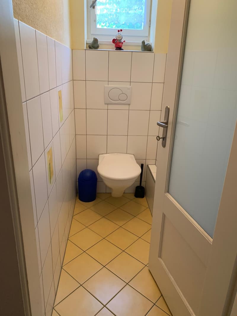 Small bathroom with white tiled walls and floor, toilet, trash bin, and toilet brush visible. Window with decorative items on sill.