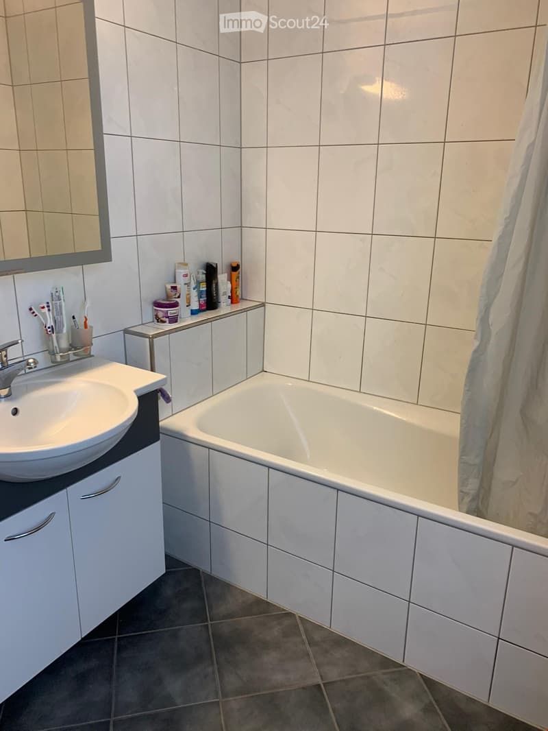 Modern bathroom with white tiles, a sink with a faucet, a bathtub, a mirror, and a shelf holding toiletries.