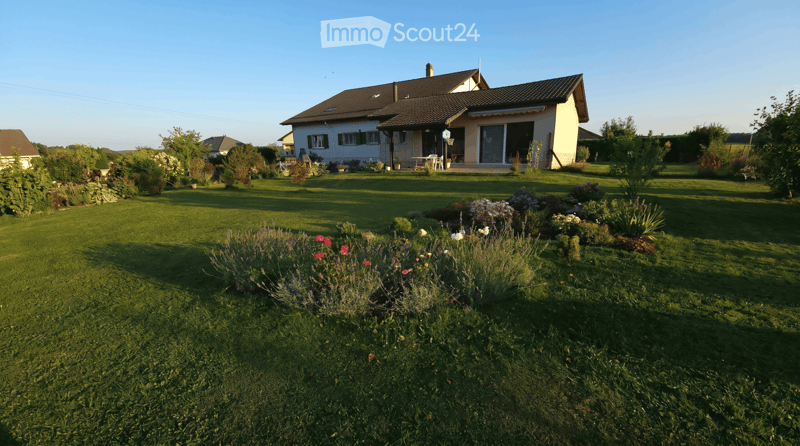 A well-maintained garden with a lawn, flower beds, and a house in the background. The house has a tiled roof, large windows, and a covered patio area with outdoor furniture.