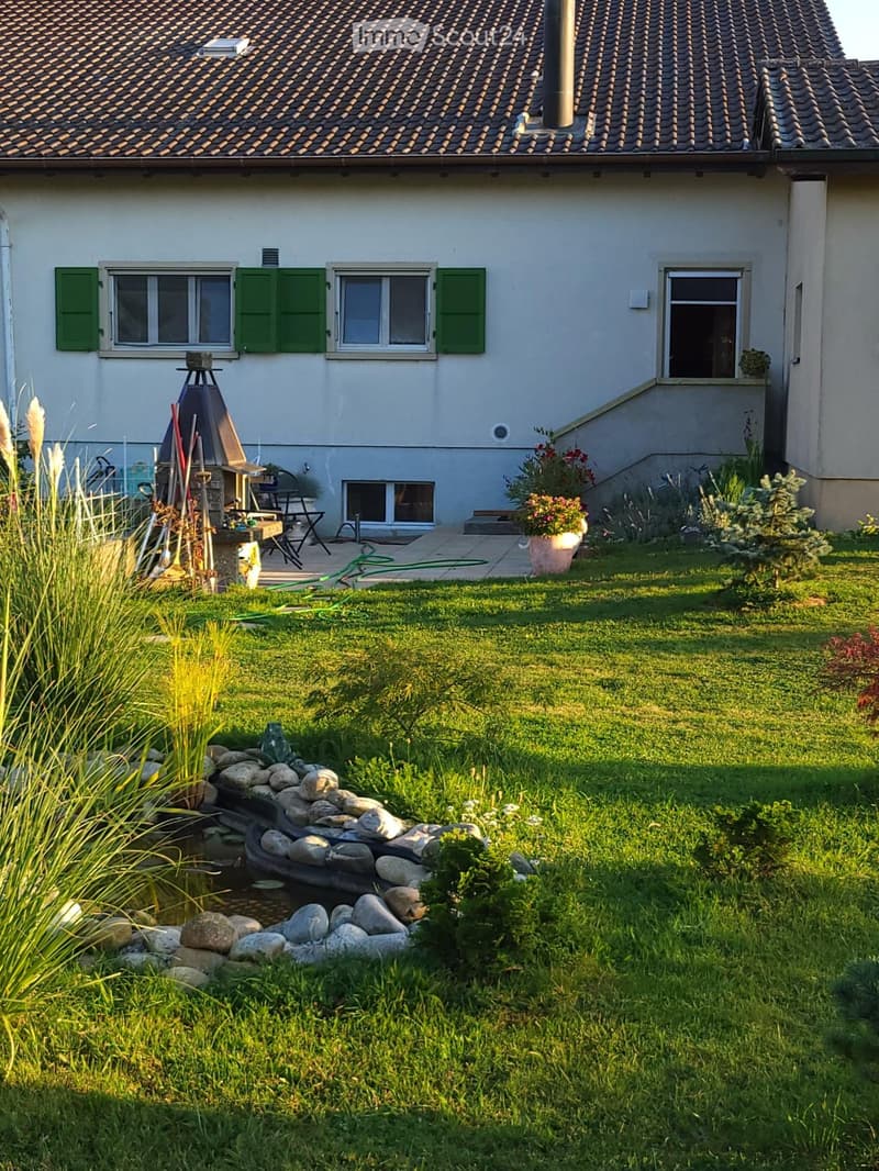 A well-maintained garden with a small pond surrounded by rocks, lush green grass, and various plants and flowers. The house features a light-colored exterior with green shutters and a small patio area with outdoor furniture.