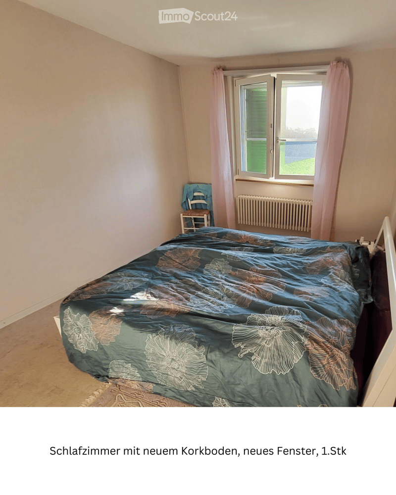 Bedroom with a large bed covered in a blue floral duvet, a small wooden chair with a blue cushion, and a radiator beneath a window with pink curtains.