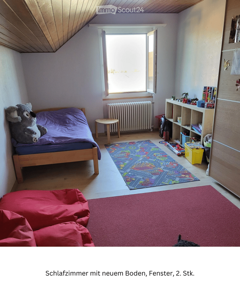 Bright bedroom with wooden floor, large window, bed with purple bedding, colorful play mat, shelf with toys, and a red bean bag sofa.