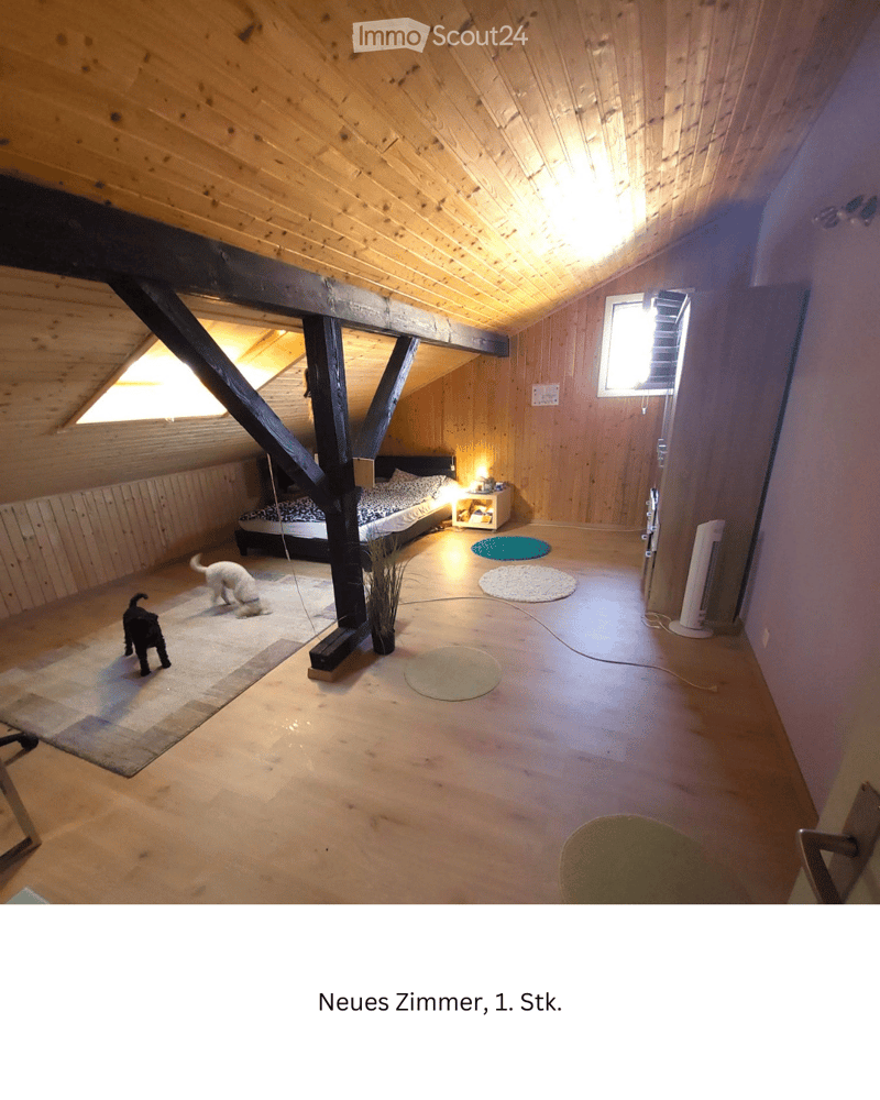 Cozy attic bedroom with wooden walls and ceiling, featuring a bed, nightstand, lamp, and fan. Two dogs are present in the room.