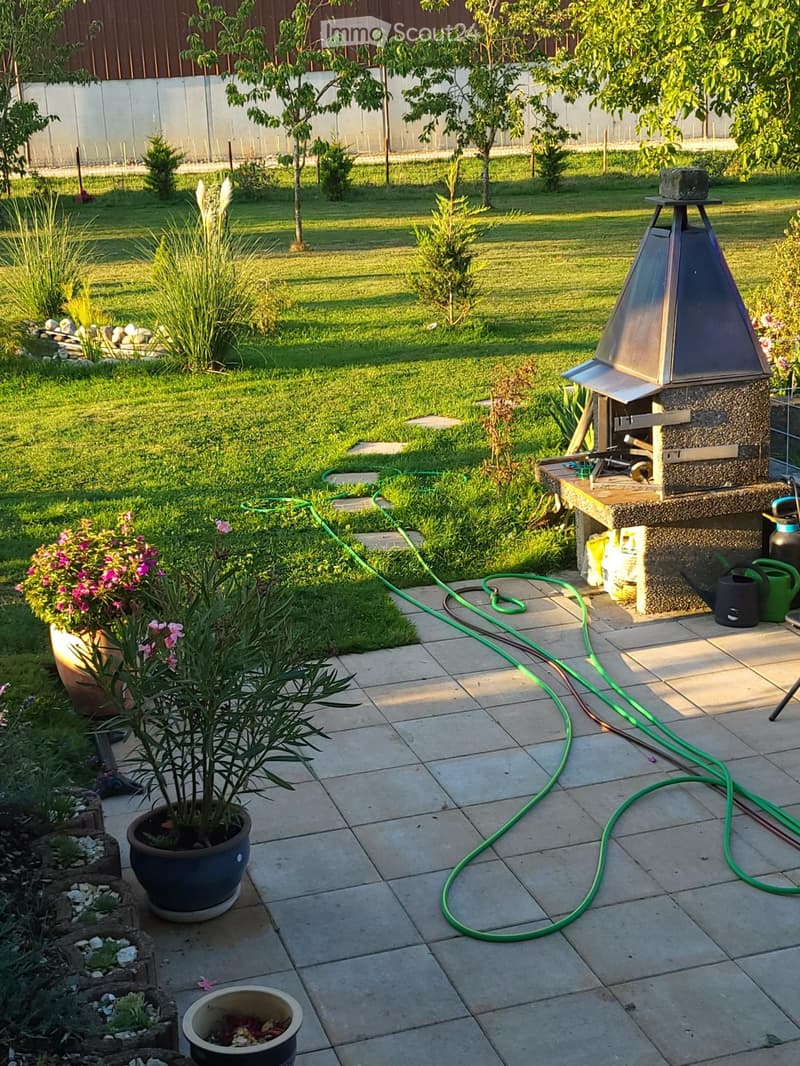 Well-maintained garden with a tiled patio, potted plants, a barbecue, and a green garden hose. The lawn is neatly trimmed, and there are various plants and shrubs around the area.