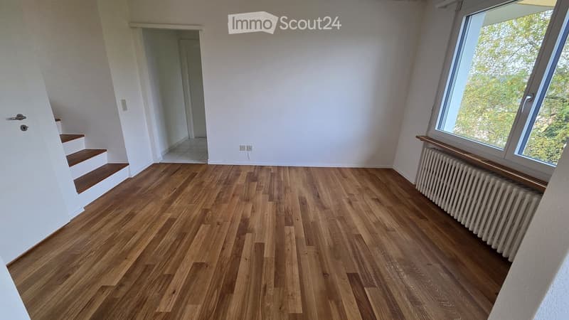 Empty room with wooden flooring, white walls, large window with view, radiator, and staircase leading upwards.