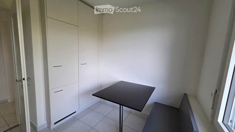 A small, modern room with a black table, a chair, and a white wardrobe with silver handles. The room has tiled flooring and white walls.