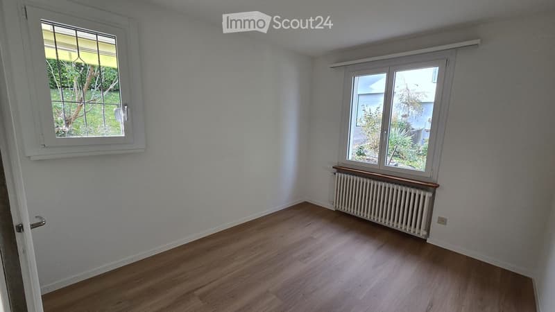 Empty room with light wooden flooring, white walls, two windows with white frames, and a radiator under one window.