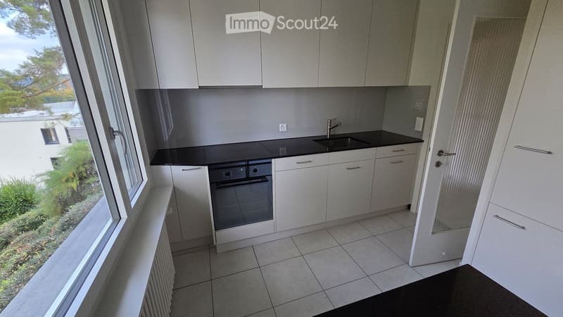 Modern kitchen with white cabinets, black countertops, a sink, and an oven. Large window offers a view of greenery outside.
