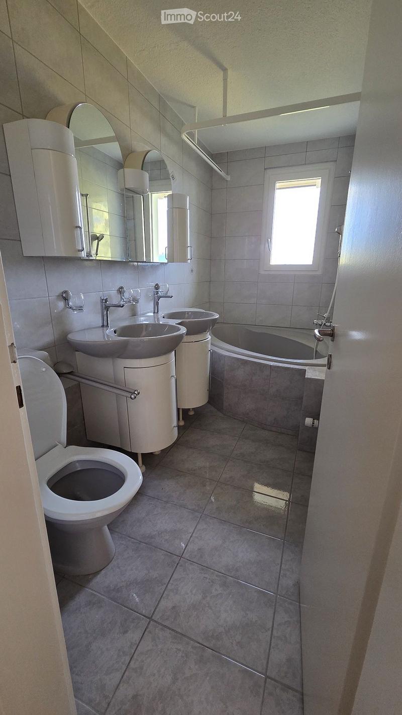 Modern bathroom with grey tiled walls and floor, featuring a toilet, double sink vanity, bathtub, and large mirror above the sinks.