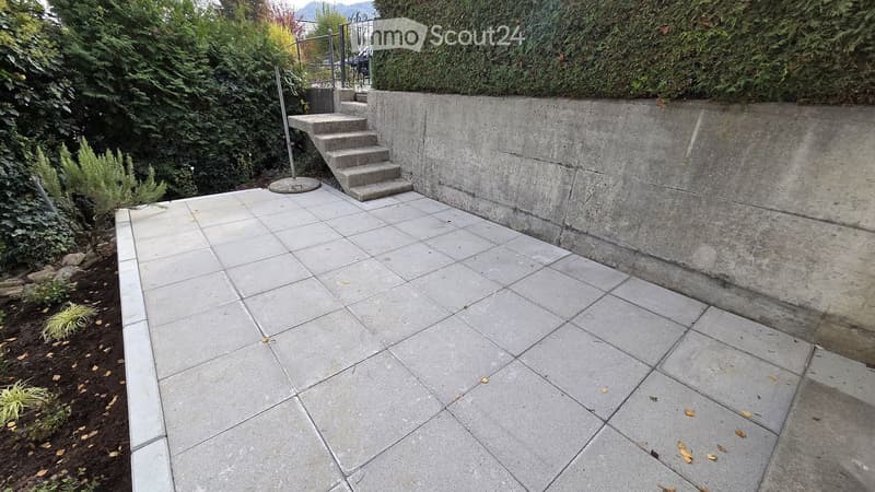 Concrete patio with large square tiles, a short concrete retaining wall, and a set of concrete steps leading up to a higher level.