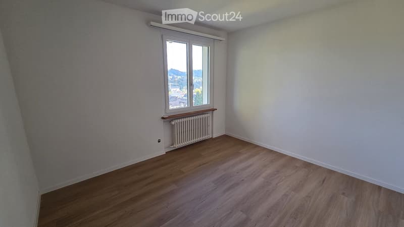 Empty room with wooden flooring, white walls, a window with a view, and a radiator below the window.