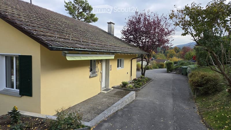 Single-story yellow house with tiled roof, green shutters, and small garden area.