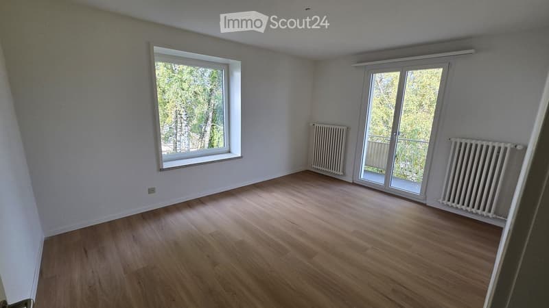 Empty room with wooden flooring, white walls, large window, sliding glass door leading to a balcony, and radiators on both sides of the door.