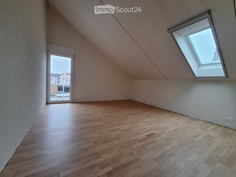 Empty attic room with wooden flooring, white walls, and a skylight window. The room features a door leading outside and a sloped ceiling.