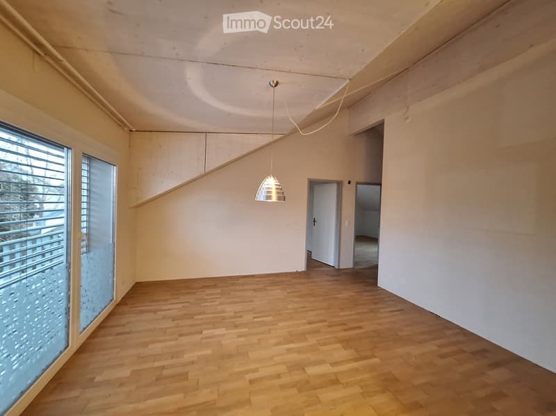 Empty room with wooden flooring, white walls, and a pendant light fixture. The room features large windows with blinds and an open doorway leading to another space.