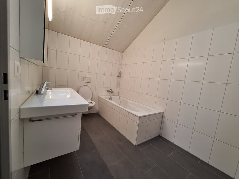 Modern bathroom with white tiles, a sink, toilet, bathtub, and a mirror. The room features a sloped ceiling typical of an attic space.
